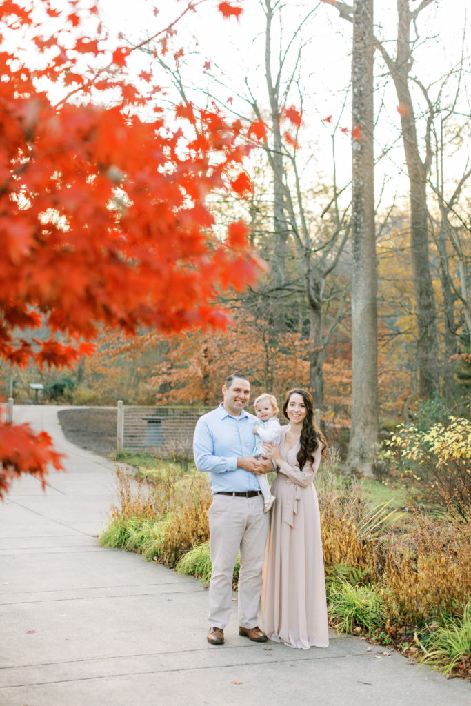 Mother, Father, And Daughter Standing In A Park During A Fall Family Photo Session.