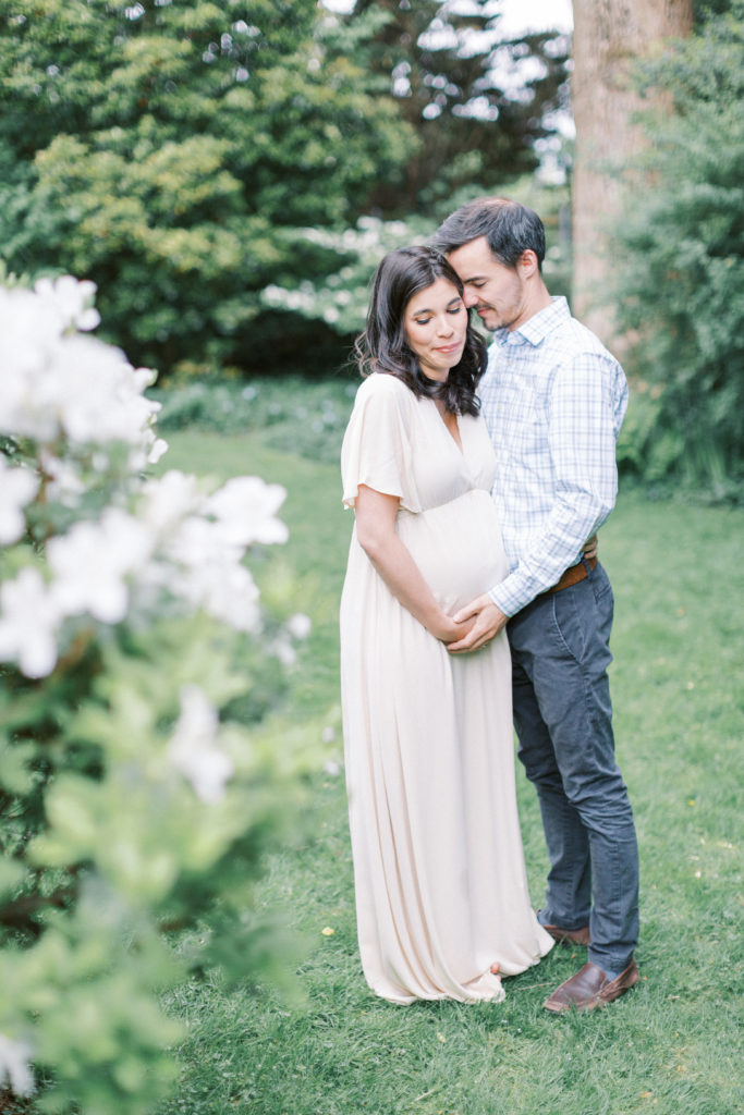 An Expecting Couple Cuddling Close Together In Spring.