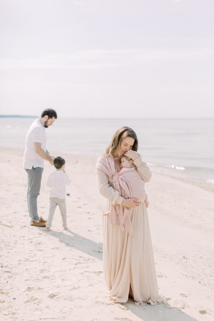 A Mother Caresses Her Young Daughter On The Beach While Her Son And Husband Play In The Background.