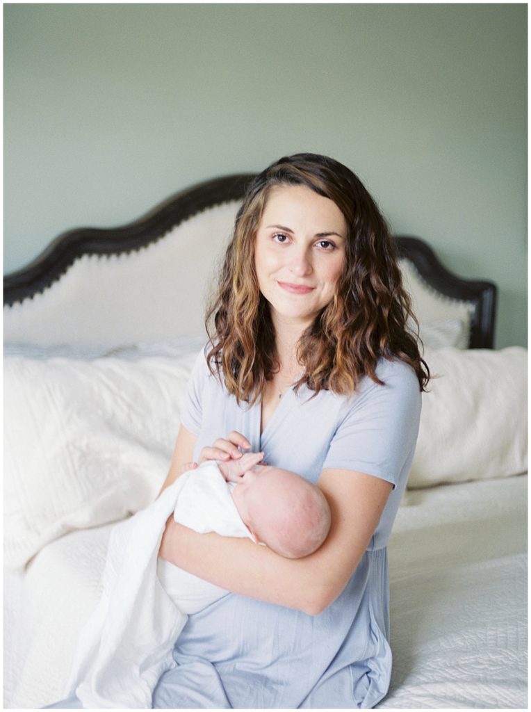 Mother Sits On The Edge Of A Bed Holding Baby During Maryland Newborn Session.