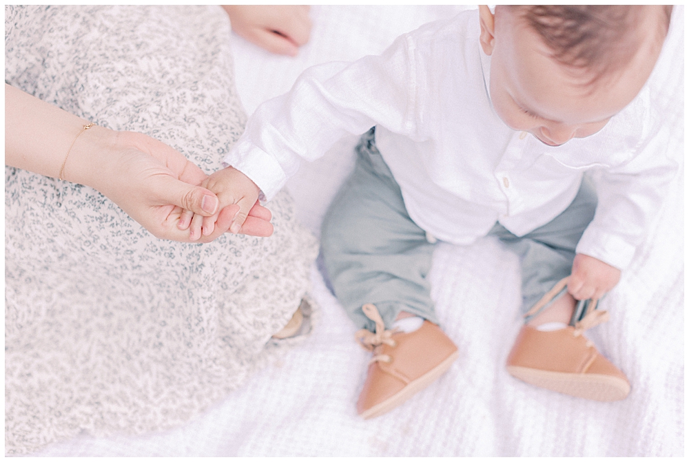 Mother Holds Baby's Hand While Sitting On A White Blanket.