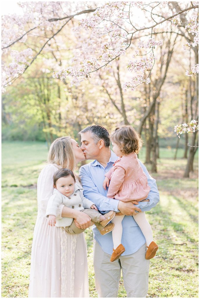 Mother And Father Kiss While Holding Their Two Young Children During A Dc Family Photography Session At The National Arboretum.