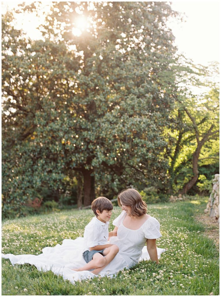 Little Boy Sits With His Mother On A White Blanket In A Field