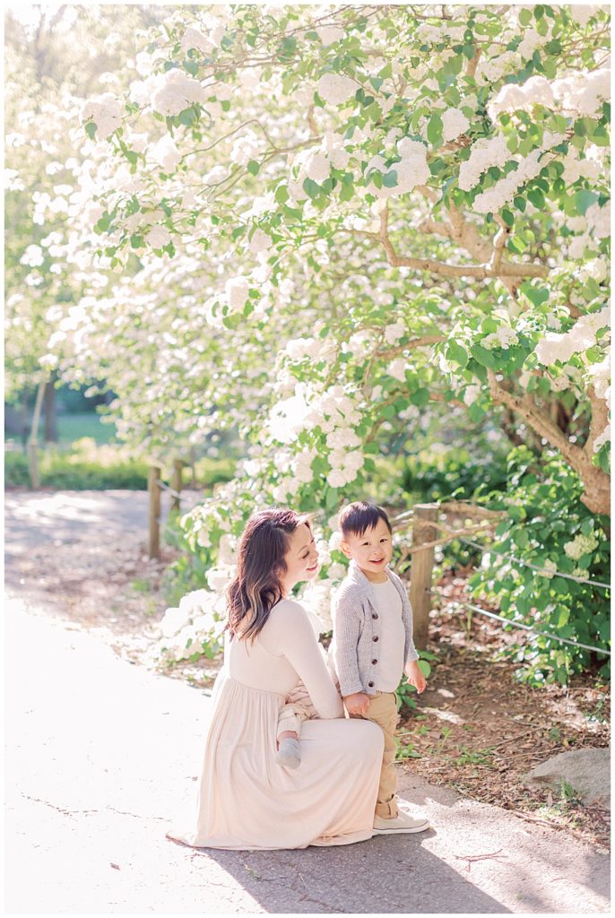 Mother Crouches Down With Son By White Flowers