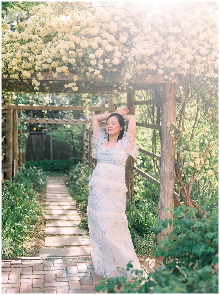 Pregnant Woman Stands With Her Hands Behind Her Head Near Floral Arch At Tudor House In Dc