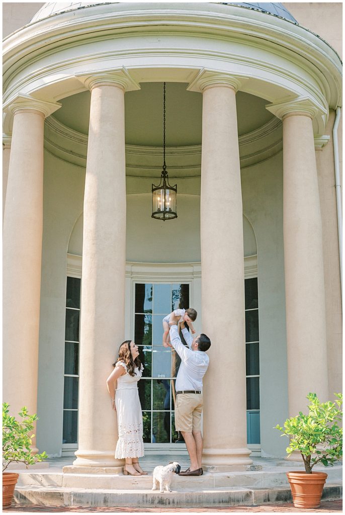 Mother And Father Stand By The Columns At Tudor Place Holding Up Their Young Son