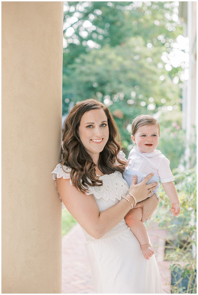 Mother Smiles While Leaning Up Against A Column Holding Her Baby Boy