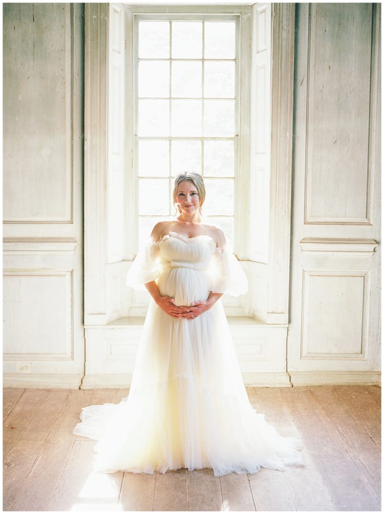 Pregnant Woman Stands With Hands Below Her Belly In Front Of A Window