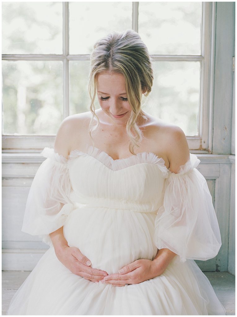 Pregnant Woman Sits With Hands Holding Belly By A Window At Salubria During Her Dc Maternity Session