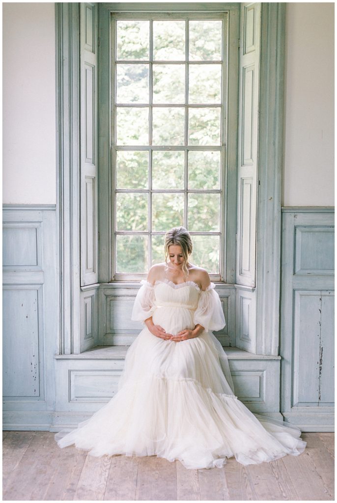 Pregnant Mother Sits In A Large Window At Salubria During Her Maternity Session