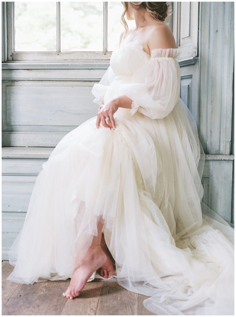 Romantic Maternity Session At Salubria | Pregnant Woman Holds Her Dress While Sitting In Window