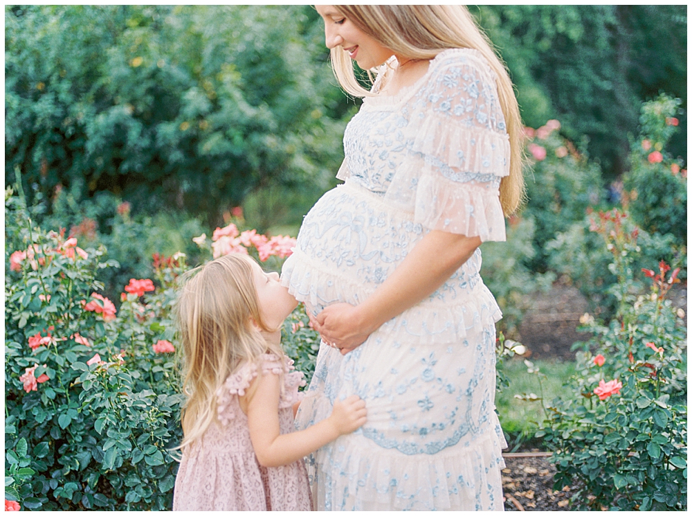 A Little Girl Kisses Her Mother's Belly During A Bon Air Rose Garden Maternity Session In Arlington, Va