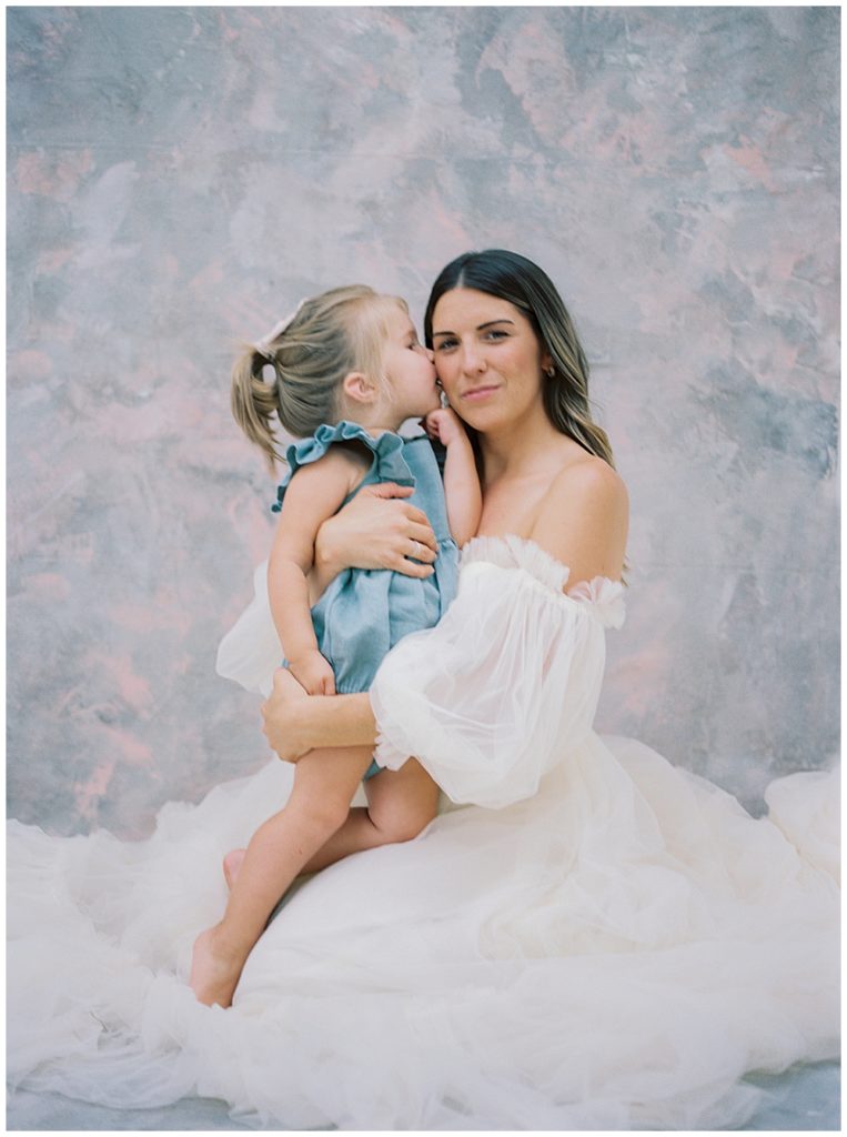 Little Girl Kisses Her Mother's Cheek During Their Studio Maternity Session