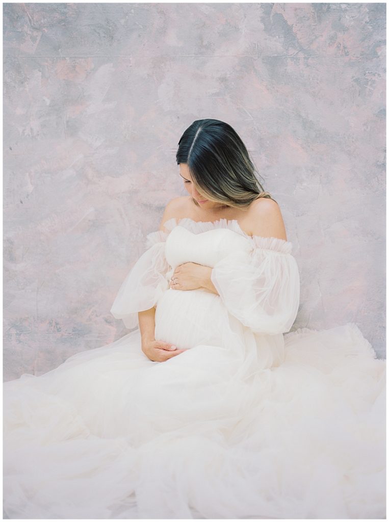 Pregnant Mother Sits With Her Hands On Her Belly Looking Down At Her Bump During Her Motherhood Studio Session In Washington Dc