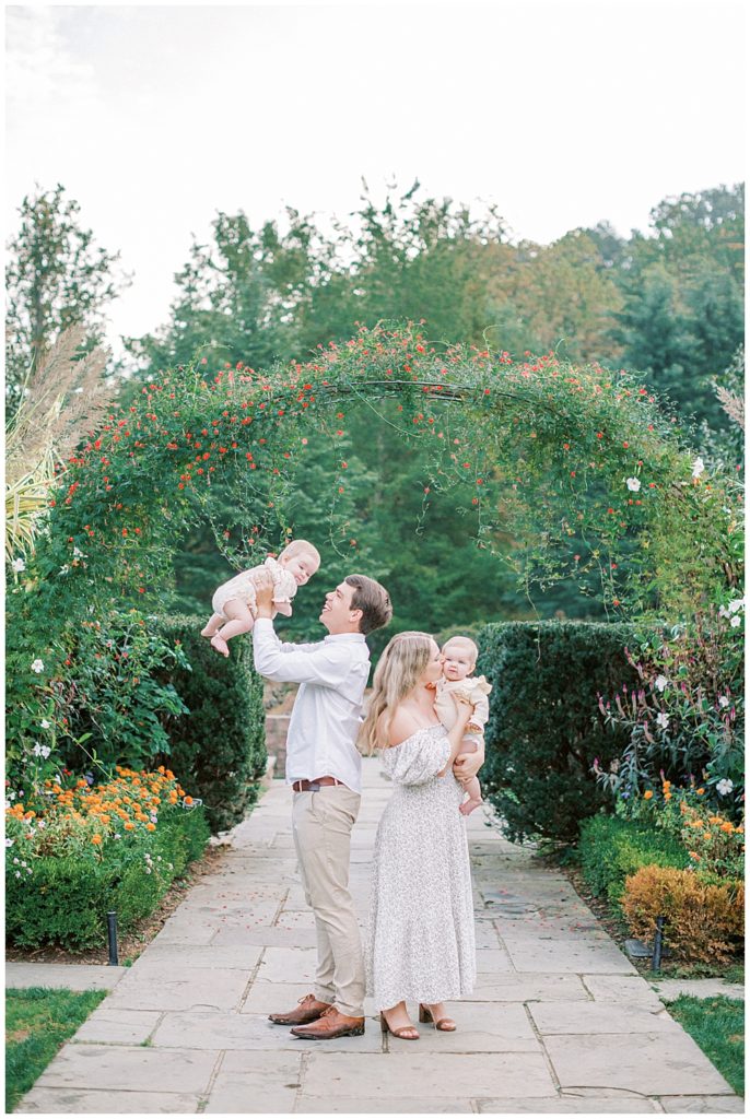 Family Stands Together With Their Babies Near Floral Arch In A Garden