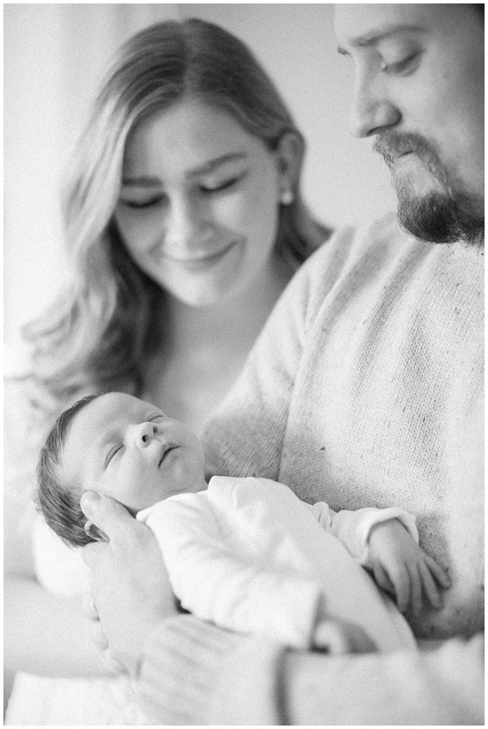 A Black And White Image Of A Father Holding His Newborn While The Mother Smiles Down At The Baby