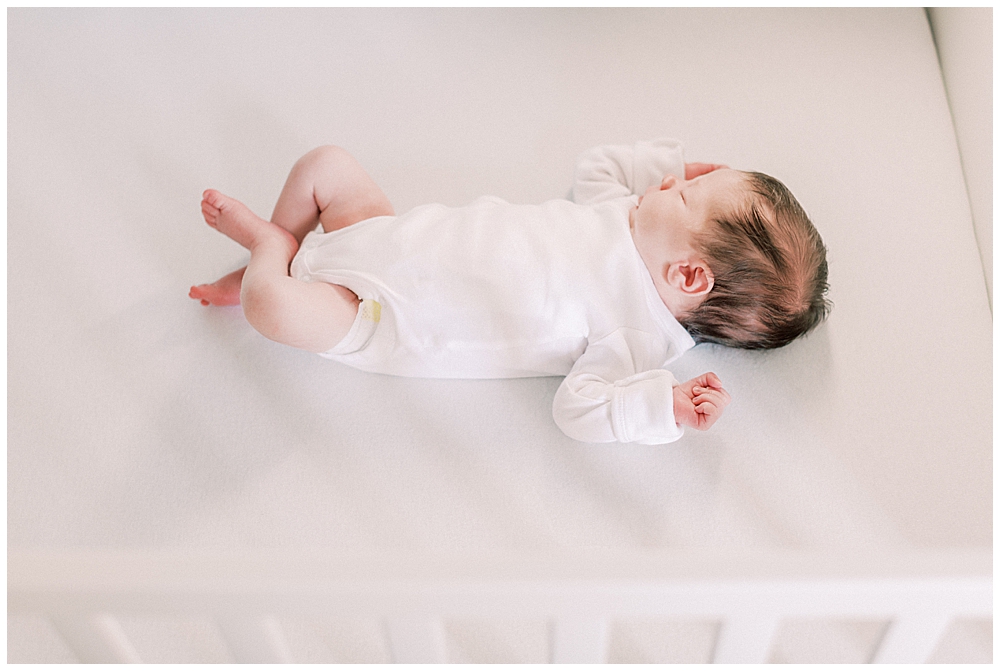 A Baby Wearing A White Onesie Lays In A White Crib During Her Newborn Session In Northern Virginia