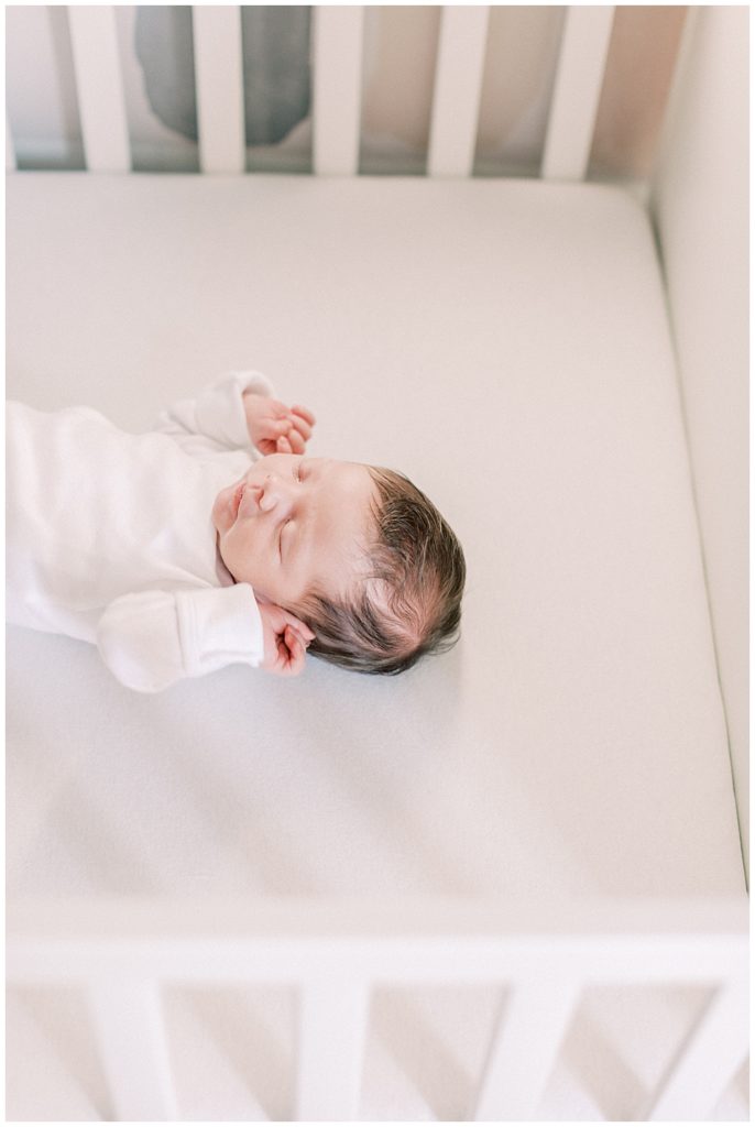 Baby Girl Lays In Her White Crib And Brings Her Hands Up To Her Head