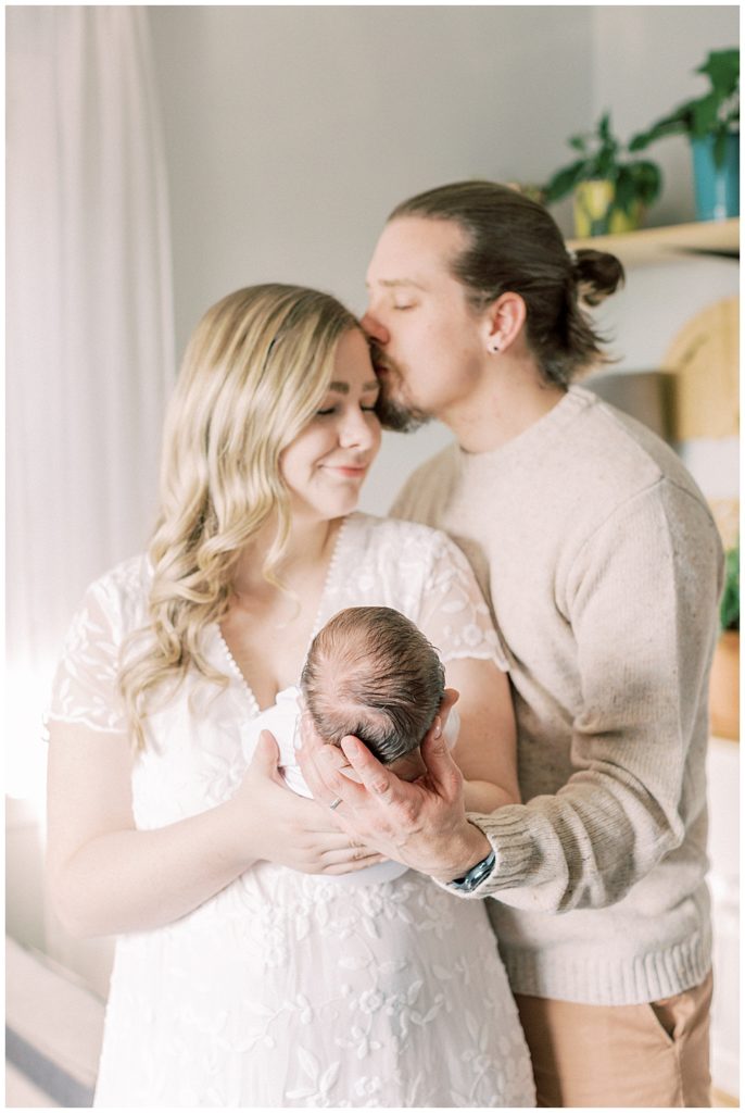 A Mother And Father Hold Their Newborn Out In Front Of Them While The Father Kisses The Mother's Forehead