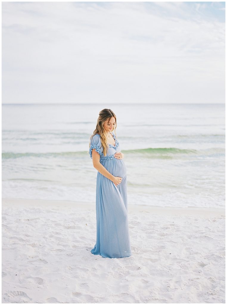 Mother Stands On The Beach In A Blue Dress With Her Hands Below Her Belly During Her Beach Maternity Session