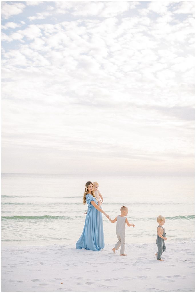 Mother Wearing A Blue Doen Dress Walks On The Beach With Her Three Sons During Her Beach Maternity Photoshoot