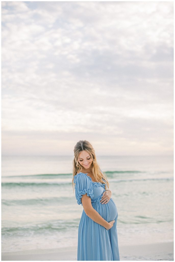 Mother Smiles Down Over Her Shoulder While Standing On The Beach Wearing A Blue Doen Dress