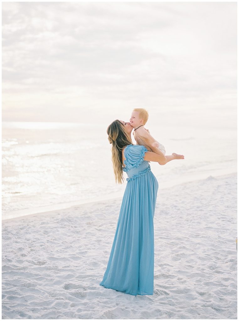 Mother Holds Up Her Son In The Air While Wearing A Blue Gown On The Beach 