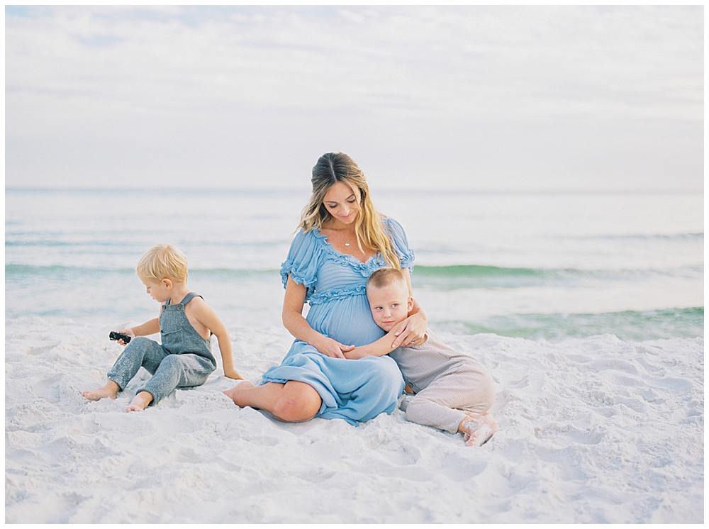 A Mother In A Blue Dress Sits On The Beach During Her Maternity Session With Her Two Young Boys, One Of Whom Is Hugging Her