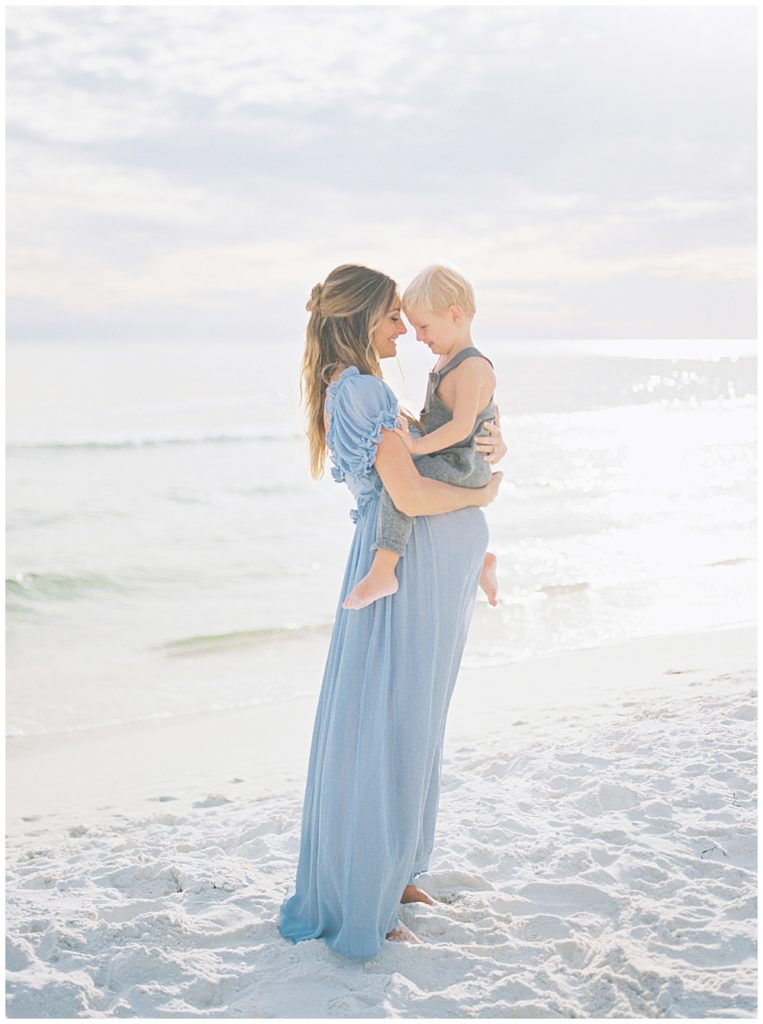 Pregnant Mother Stands On The Beach Wearing A Blue Dress And Holding Her Son During Her Beach Maternity Photoshoot