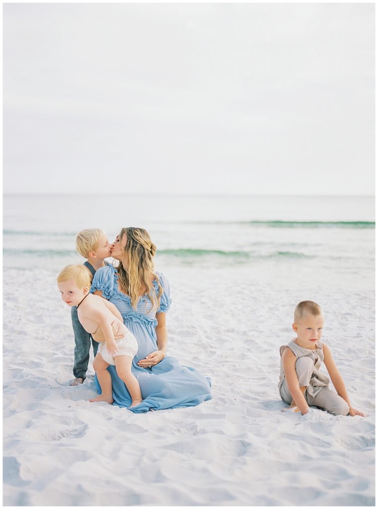 Mother Kneels On The Beach And Kisses Her Young Son Behind Her While Her Other Two Young Sons Play In The Sand