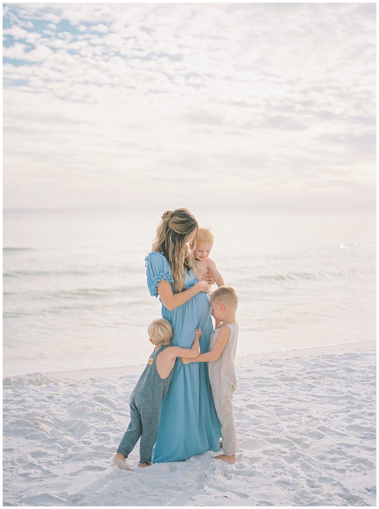 Mother Stands On The Beach While Her Three Young Sons Give Her A Hug