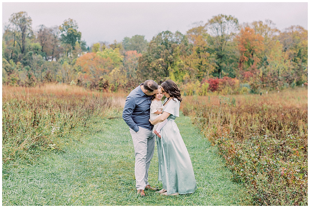 Mother And Father Hold Their Toddler Daughter And Lean Into Her At The Howard County Conservancy During Their Family Photo Session By Maryland Family Photographer Marie Elizabeth Photography