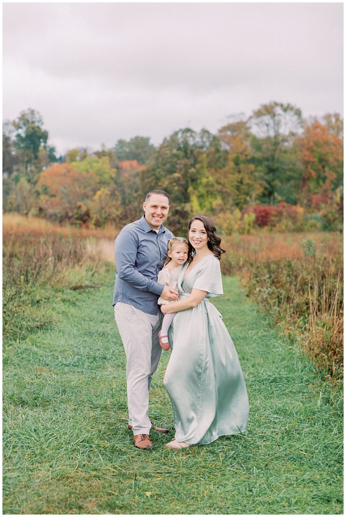 Mother And Father Hold Their Toddler Daughter While Standing In A Clearing At The Howard County Conservancy During Their Fall Family Photos By Maryland Family Photographer Marie Elizabeth Photography