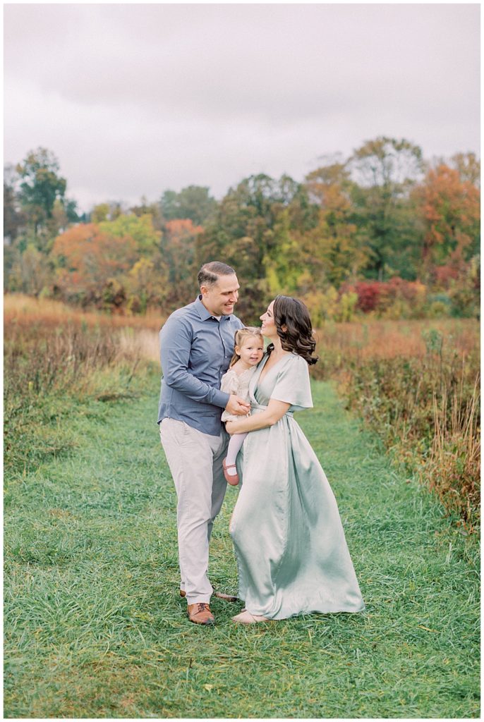 Mother And Father Hold Their Toddler Daughter And Look At One Another During Their Photo Session At The Howard County Conservancy