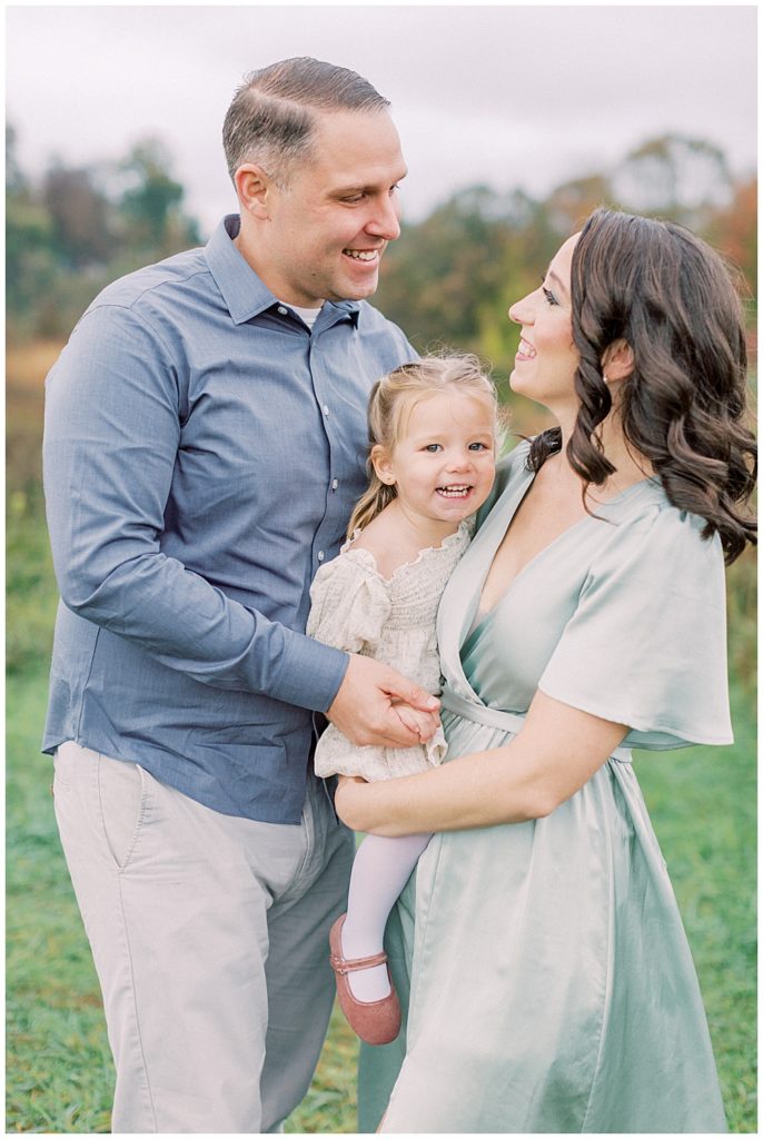 Mother And Father Smile At One Another While Holding Their Little Girl During Their Maryland Family Photo Session