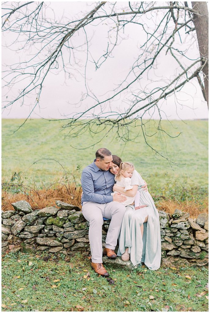 Mother Holds Her Daughter Nand Rests Her Head On Her Husband's Shoulders While Sitting On The Stone Fence At The Howard County Conservancy