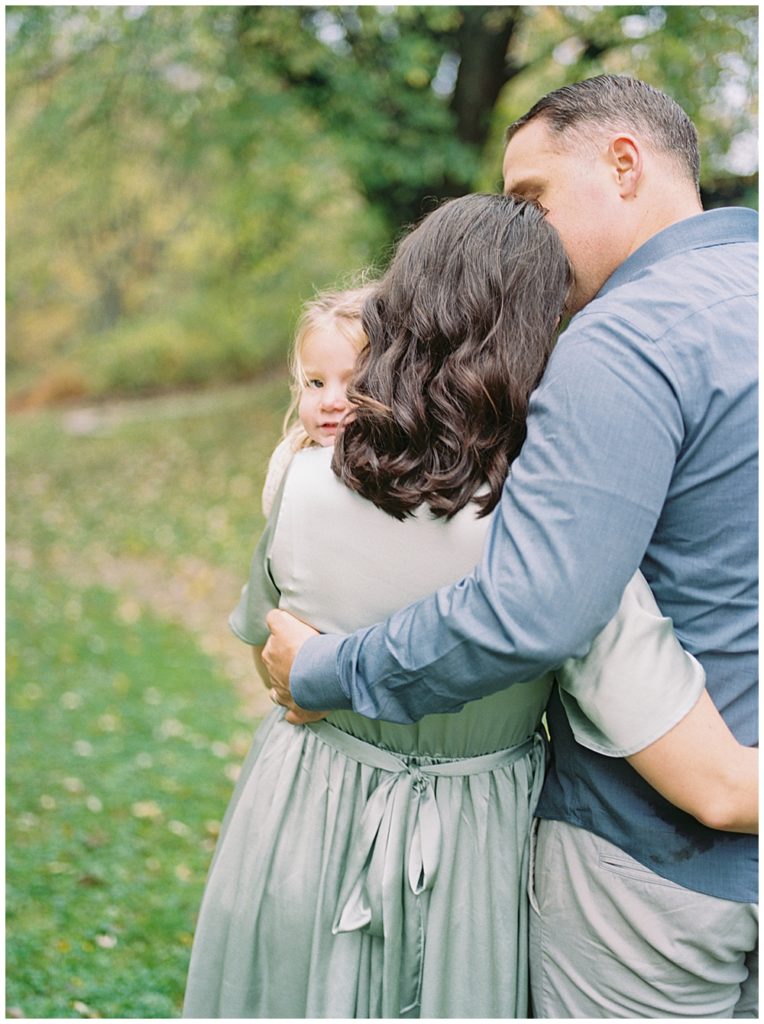 Little Girl Peaks Behind Her Mother's Shoulder While Her Parents Hold Her