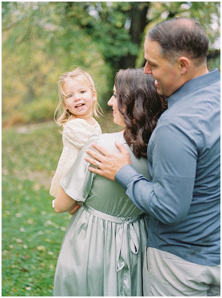 Little Girl Smiles At The Camera While Her Parents Look Adoringly At Her