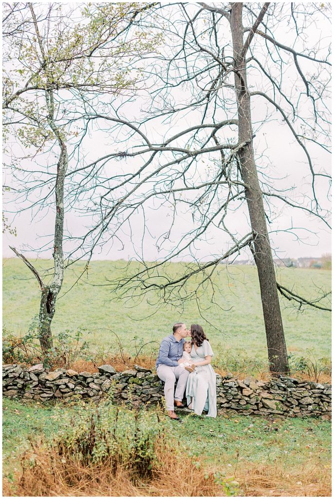 Mother, Father, And Toddler Daughter Sit On The Stone Wall At The Howard County Conservancy During Their Family Photo Session