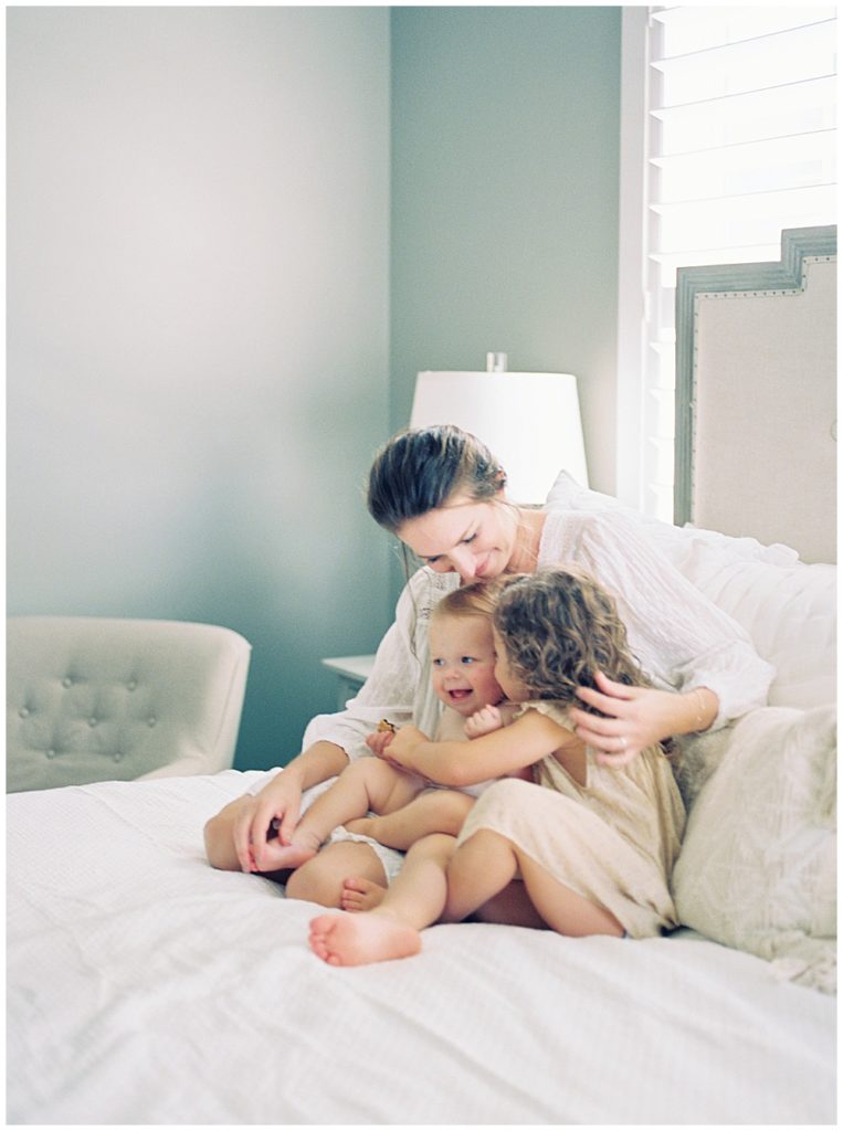 A Mother Sits On The Bed With Her Two Young Daughters And Caresses Them Close To Her During Her Lifestyle Family Session