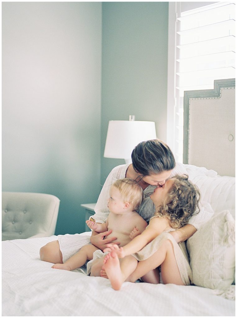 A Mother Sits On The Bed With Her Toddler Daughter And Infant Daughter And Leans In To Give Her Toddler A Kiss