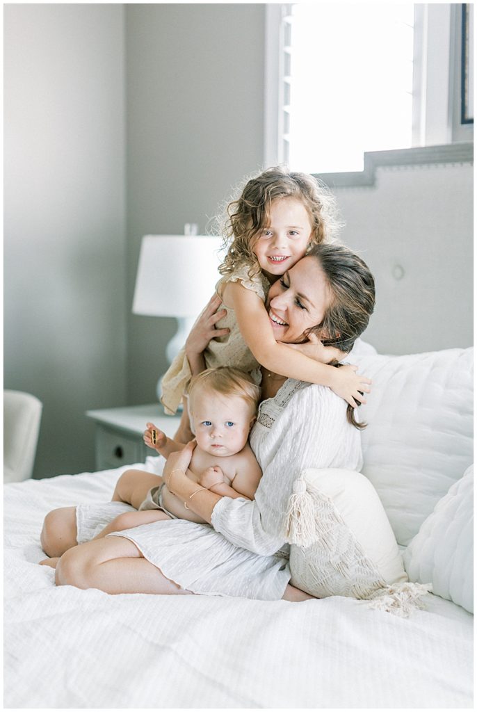 A Toddler Girl Stands On The Bed And Hugs Her Mother Who Is Sitting With Her Infant Daughter On Her Lap