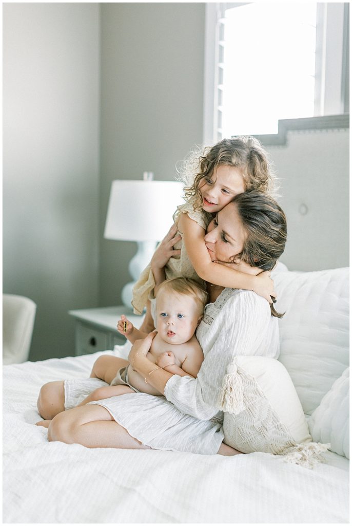 A Little Girl Stands On The Bed An Hugs Her Mother While Her Mother Sits And Holds Her Other Daughter On Her Lap During Their Family Photo Session