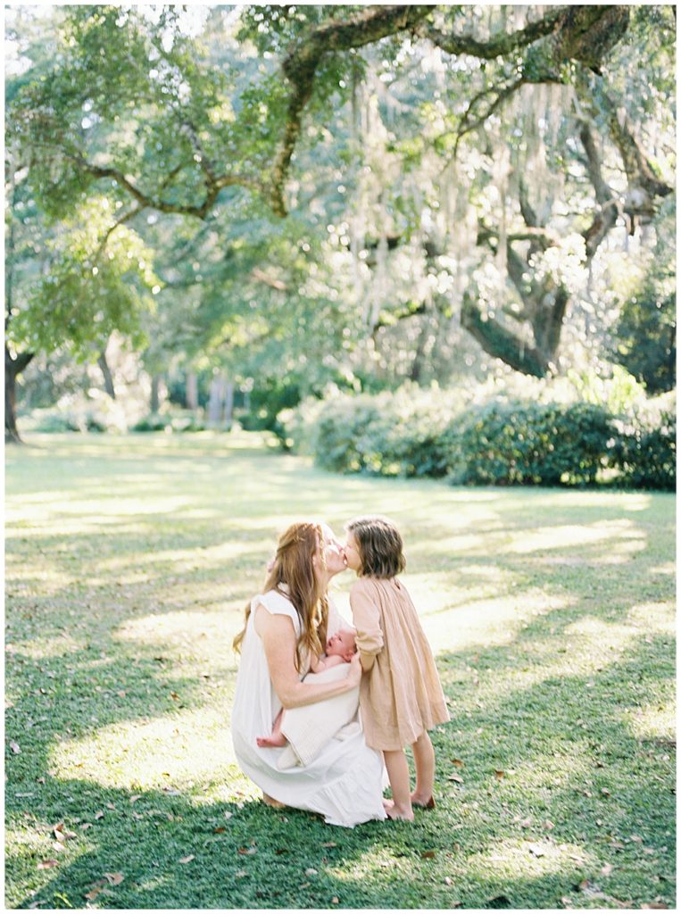 A Mother Kneels Down Outside Holding Her Newborn Baby And Kisses Her Toddler During Their Outdoor Newborn Session