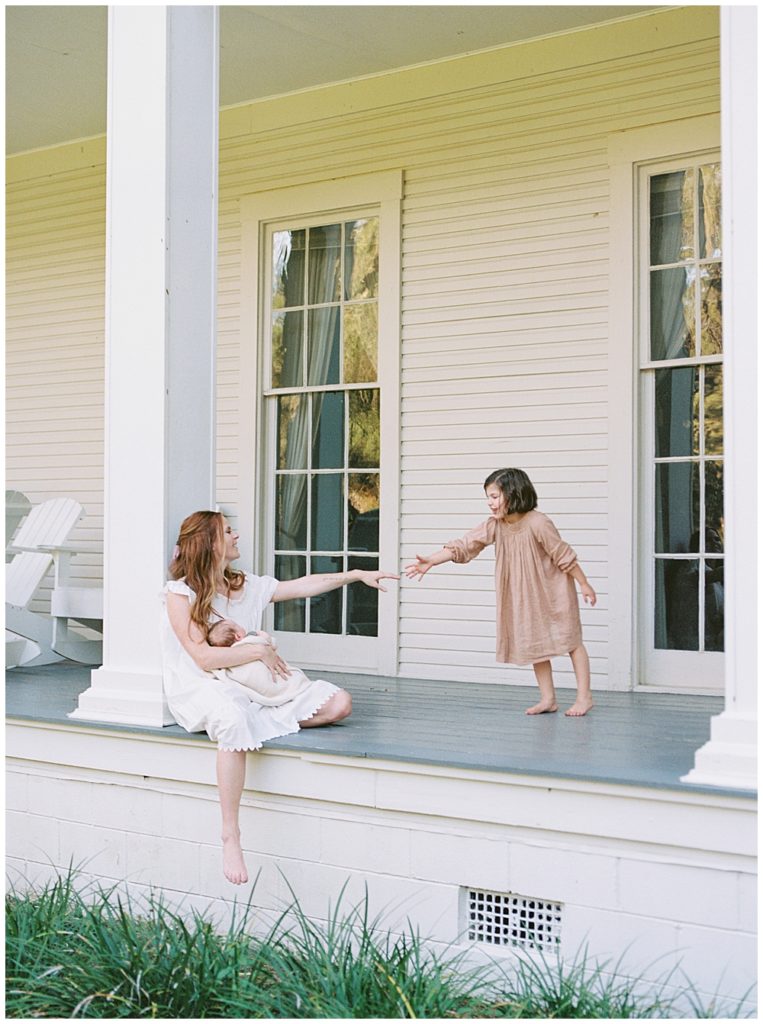 A Mother Sits On The Porch Holding Her Newborn And Takes Her Hand To Reach For Her Daughter Who Is Reaching Her Hand Towards Her