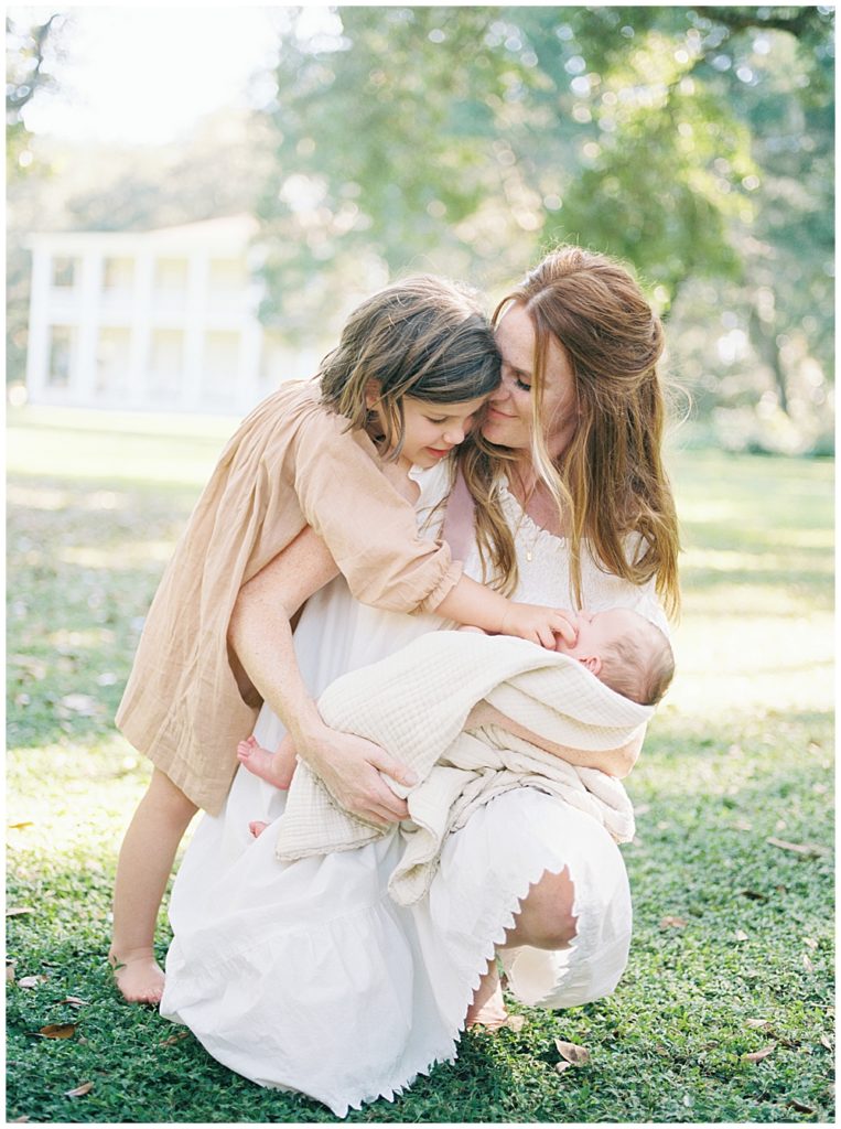 A New Mother Crouches Down While Holding Her Newborn Baby While Her Young Daughter Leans Over Her Caressing The Newborn