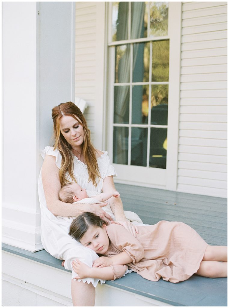 A Little Girl Lays On Her Mother's Lap While Her Mother Holds Her Newborn Baby On The Porch