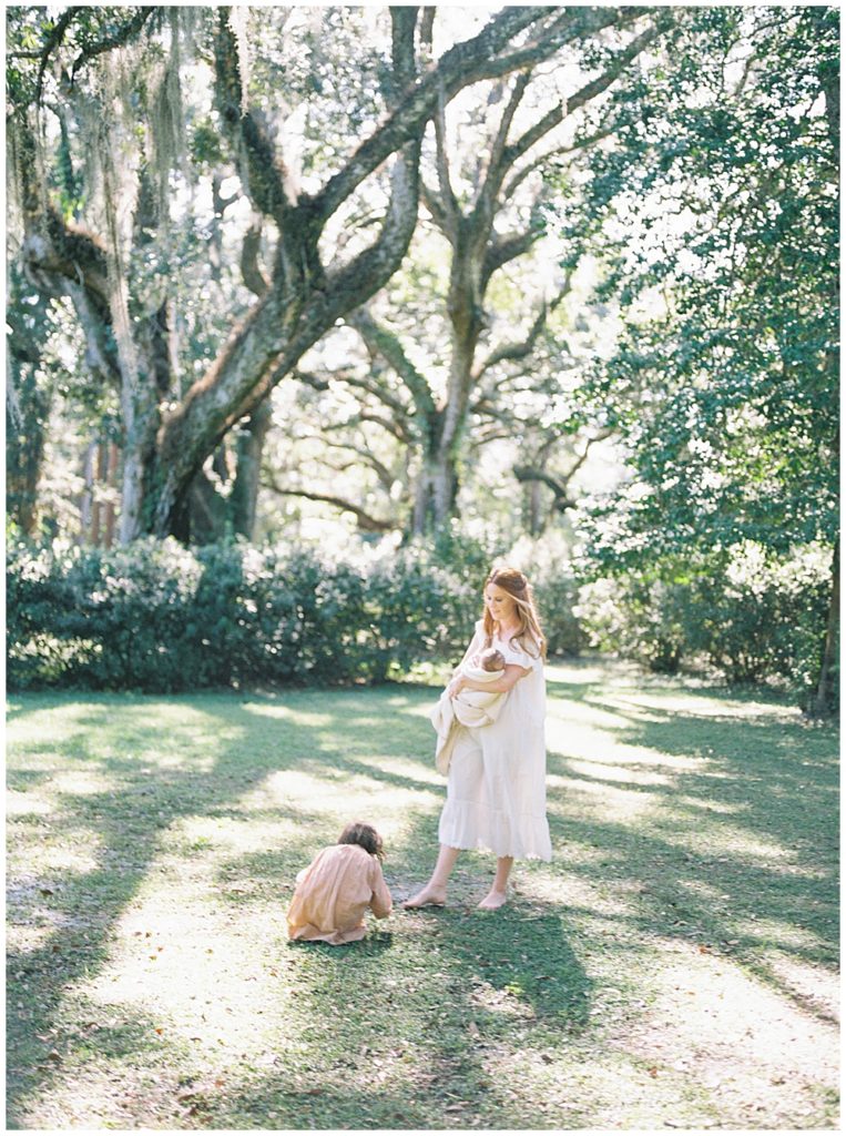 A Red Haired Mother Holds Her Newborn Baby Outside During Her Outdoor Newborn Session While Her Daughter Kneels Over And Plays In The Dirt In Front Of Her