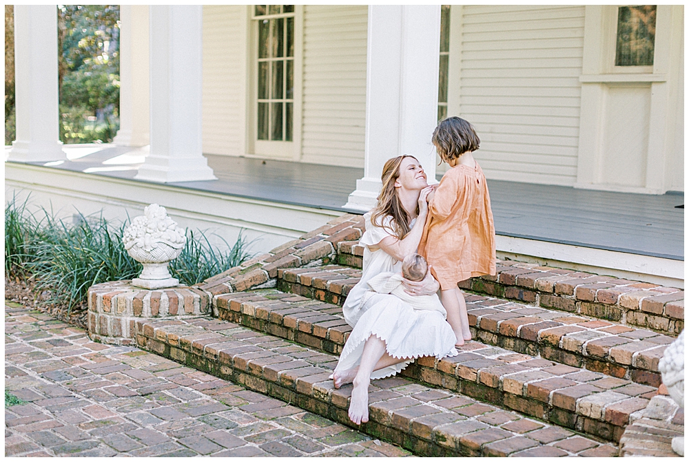 A Little Girl Stands On Brick Steps With Her Mother Who Is Sitting And Holding A Baby. The Two Look Lovingly At One Another.