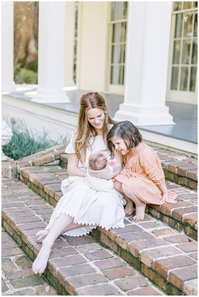 A Little Girl Sits On The Steps With Her Mother And Baby Brother And Leans Over To Look At Her Baby Brother